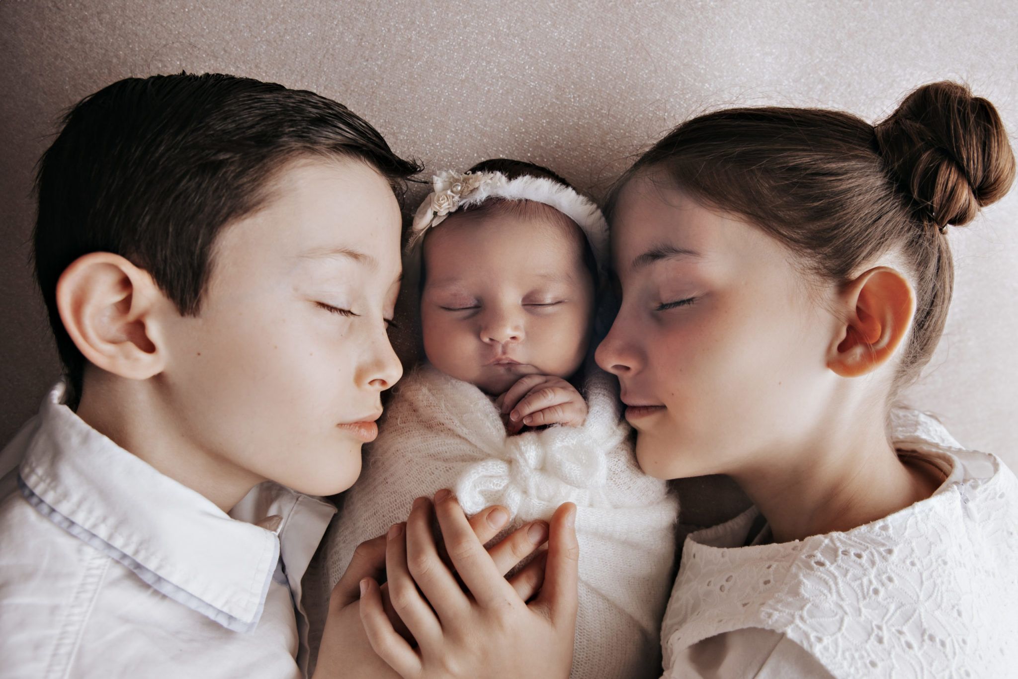 Portrait de fratrie : un garçon et une fille serrant tendrement leur petite sœur nouveau-née endormie, les yeux fermés, dans une ambiance douce et apaisante