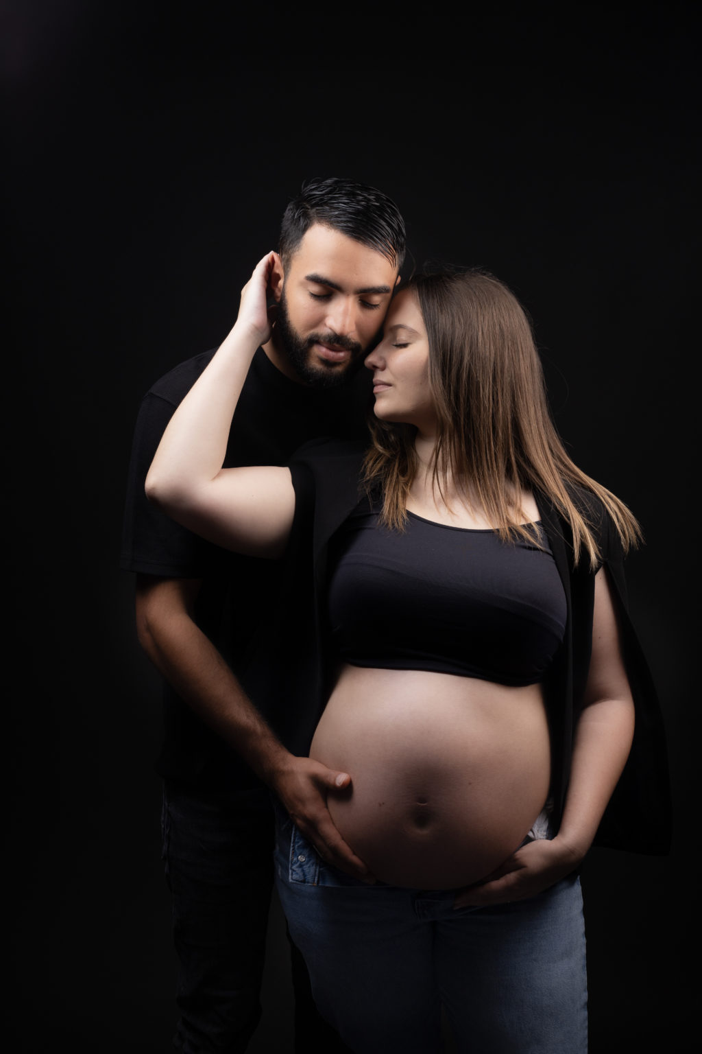 Couple en séance grossesse, en tenue noire sur fond sombre, la future maman tenant son ventre et regardant tendrement son partenaire