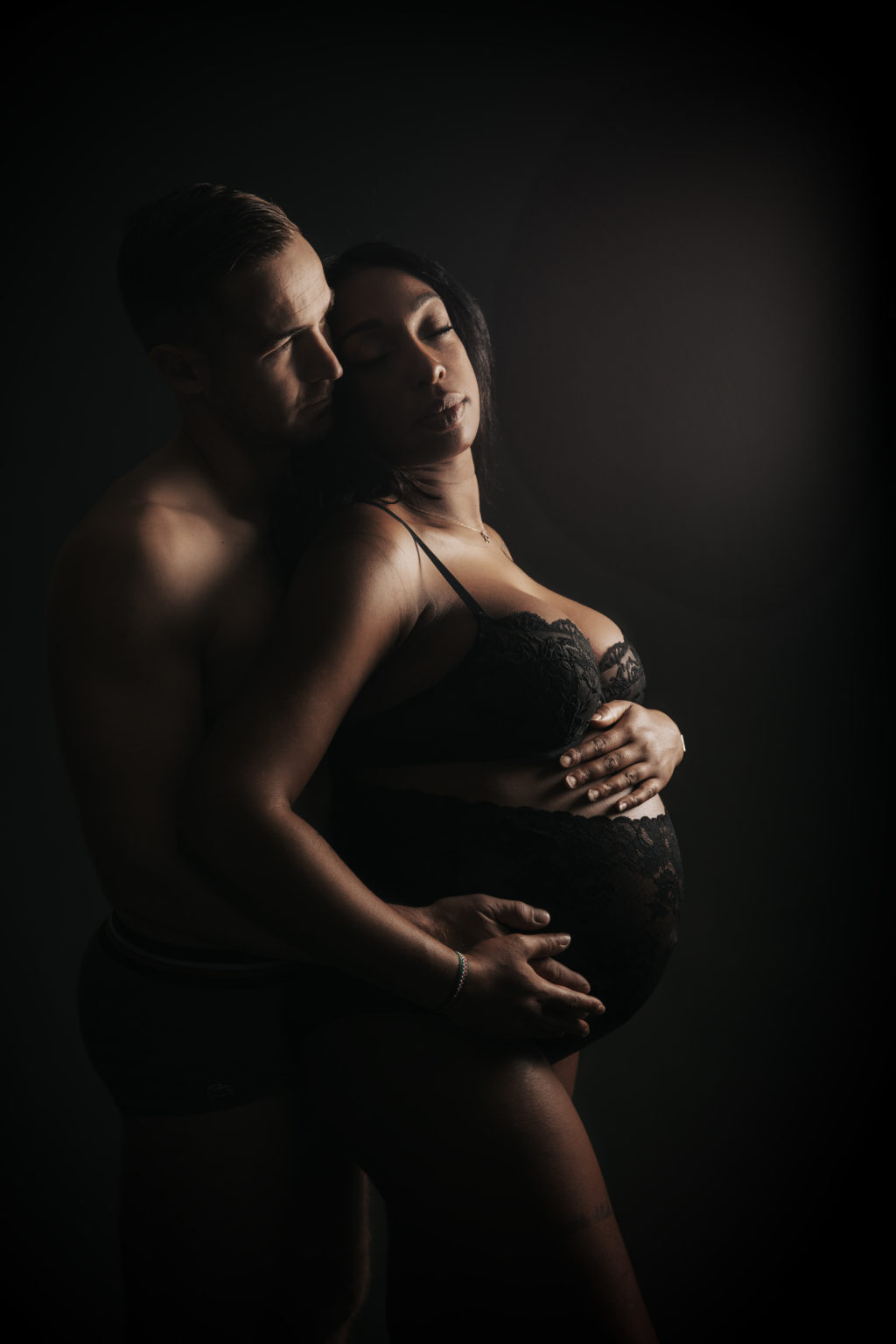 Femme enceinte en lingerie noire en lumière tamisée, tendrement enlacée par son compagnon dans un portrait intimiste et artistique