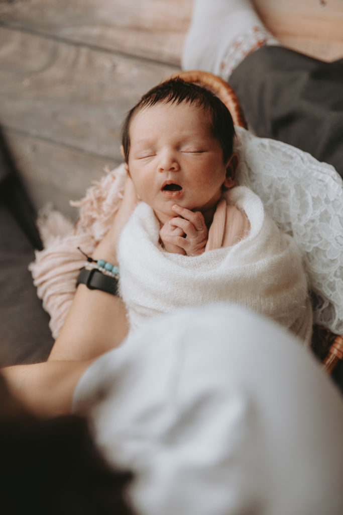 Photo en coulisses d’une séance nouveau-né : bébé emmailloté et endormi dans les bras du photographe, en ambiance naturelle et cocooning