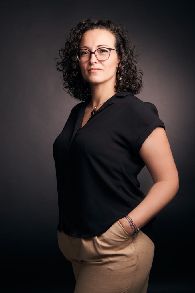 Portrait professionnel de Cécile Muselet, photographe, debout de face, main dans la poche, regard déterminé et bienveillant, sur fond studio foncé