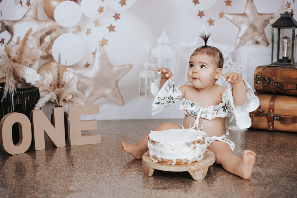 Bébé assise devant un gâteau d’anniversaire, les mains levées, dans un décor festif avec étoiles dorées