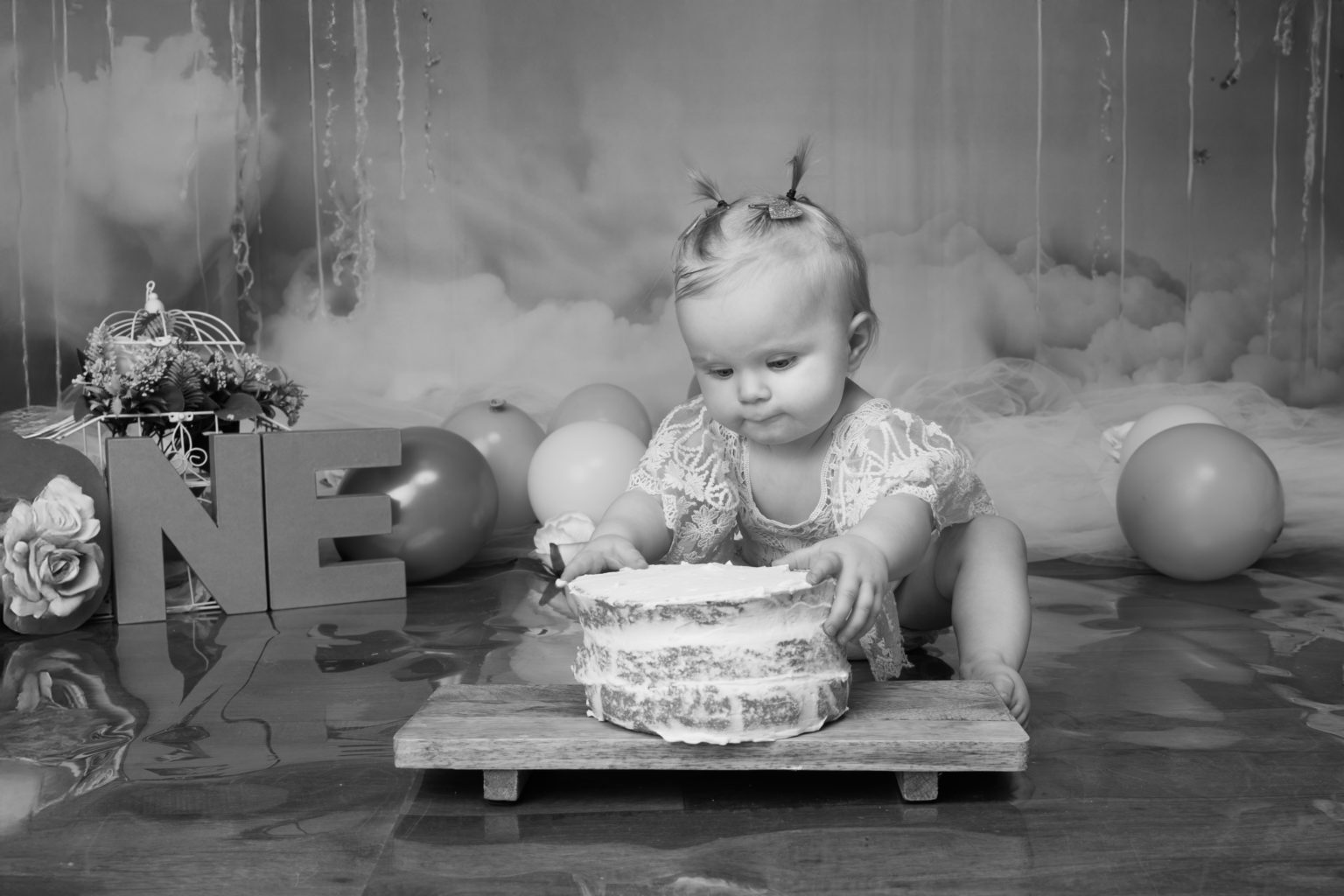Bébé avec deux petites couettes jouant avec son gâteau d'anniversaire – séance smash the cake en noir et blanc