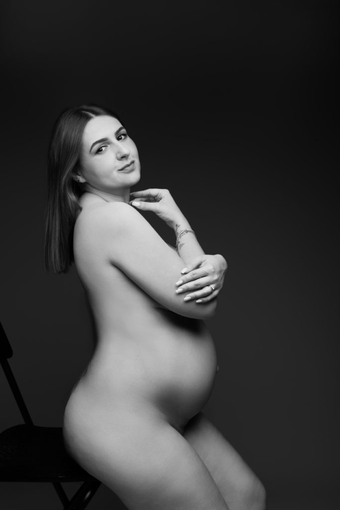 Portrait en noir et blanc d’une femme enceinte nue, assise sur une chaise, regard vers l’objectif, bras croisés avec élégance sur son buste