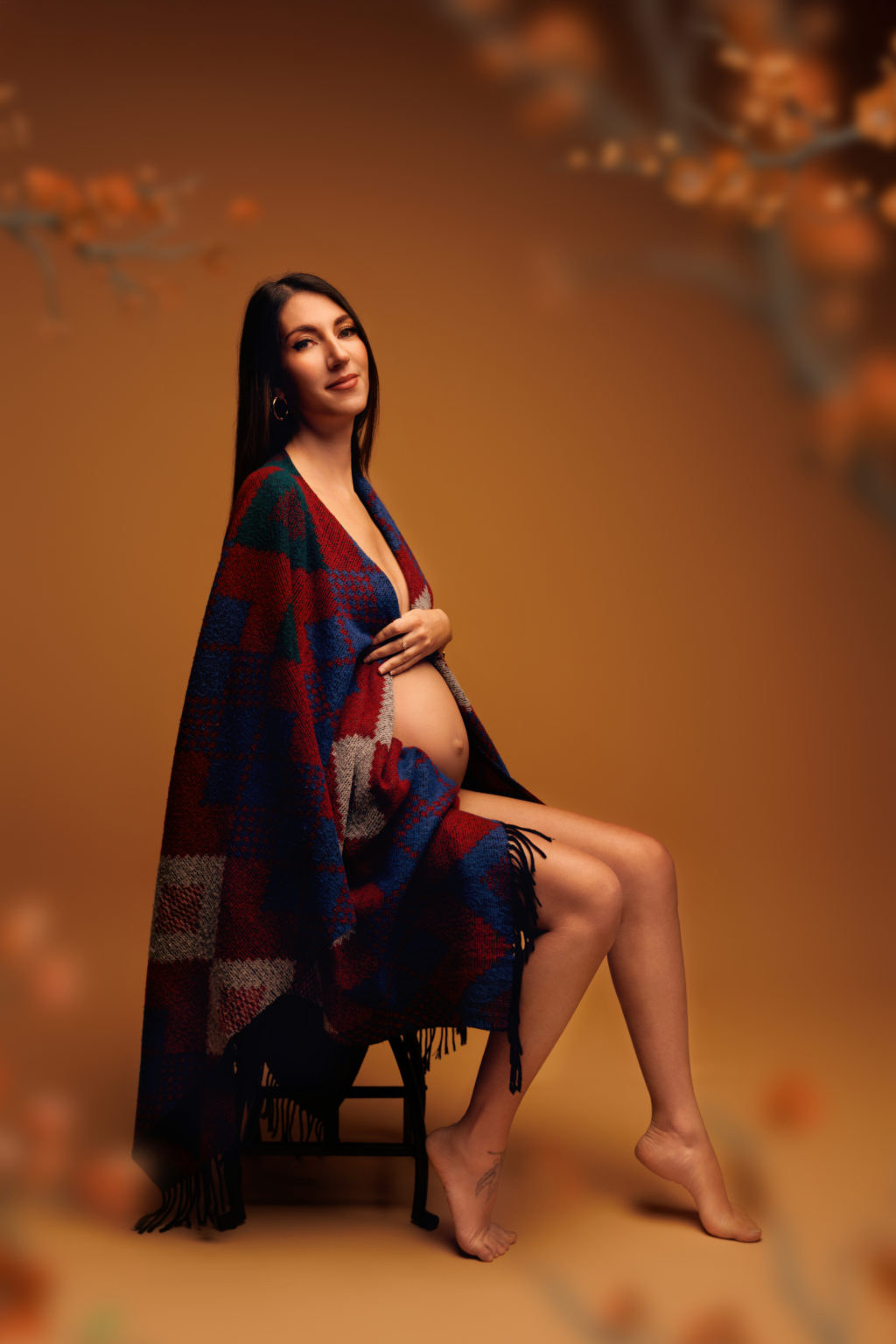 Femme enceinte assise, enveloppée dans un plaid coloré, posant sereinement sur fond ocre avec ambiance automnale en studio