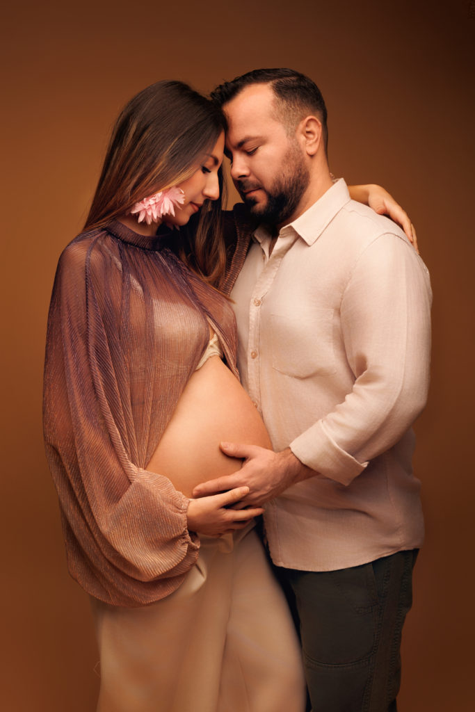 Femme enceinte et son compagnon en studio, front contre front, mains posées sur le ventre rond, dans des tons chauds et doux