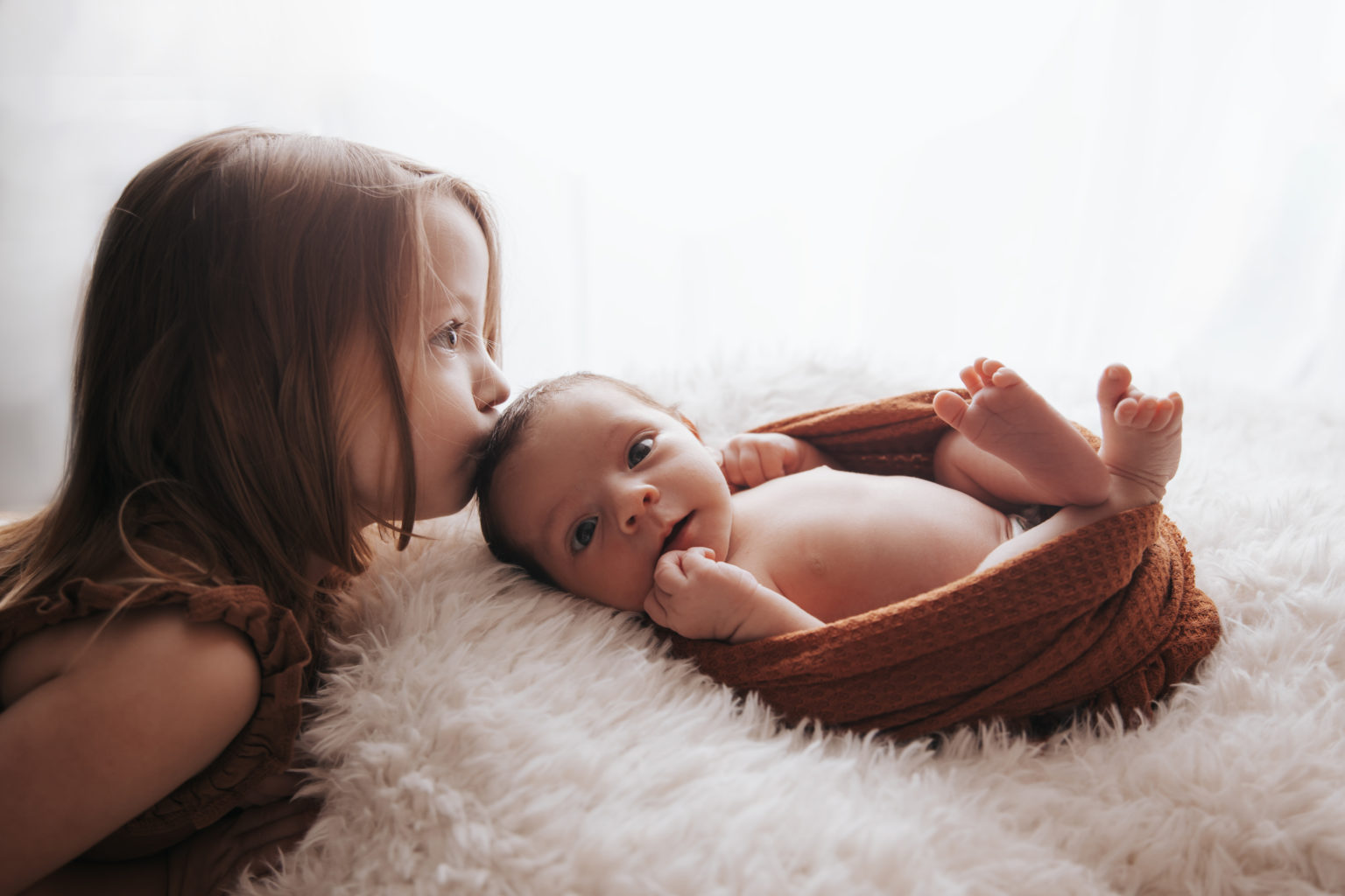 Petite fille embrassant tendrement son bébé frère allongé sur un plaid blanc.