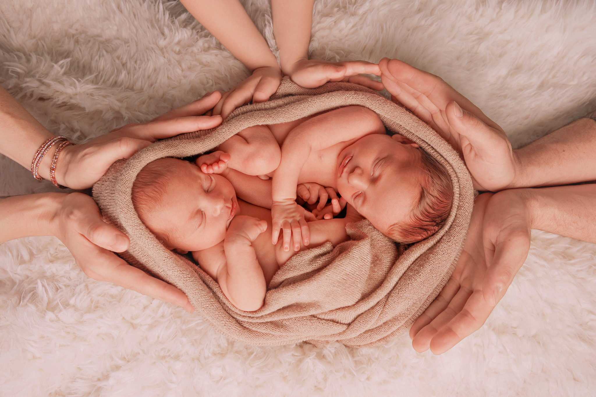Deux nouveau-nés jumeaux endormis, lovés ensemble dans une couverture beige, entourés des mains protectrices de leur famille.
