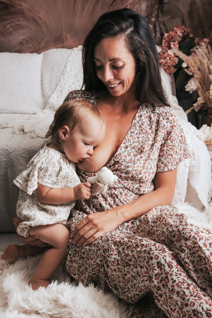 Maman assise en robe fleurie allaitant tendrement sa petite fille, qui tient une peluche et est blottie contre elle, dans un décor doux et chaleureux