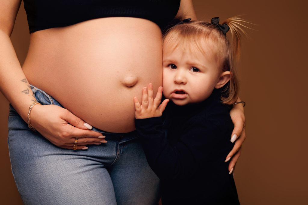 Petite fille tenant le ventre rond de sa maman enceinte, posant contre lui avec émotion sur fond chaud en studio