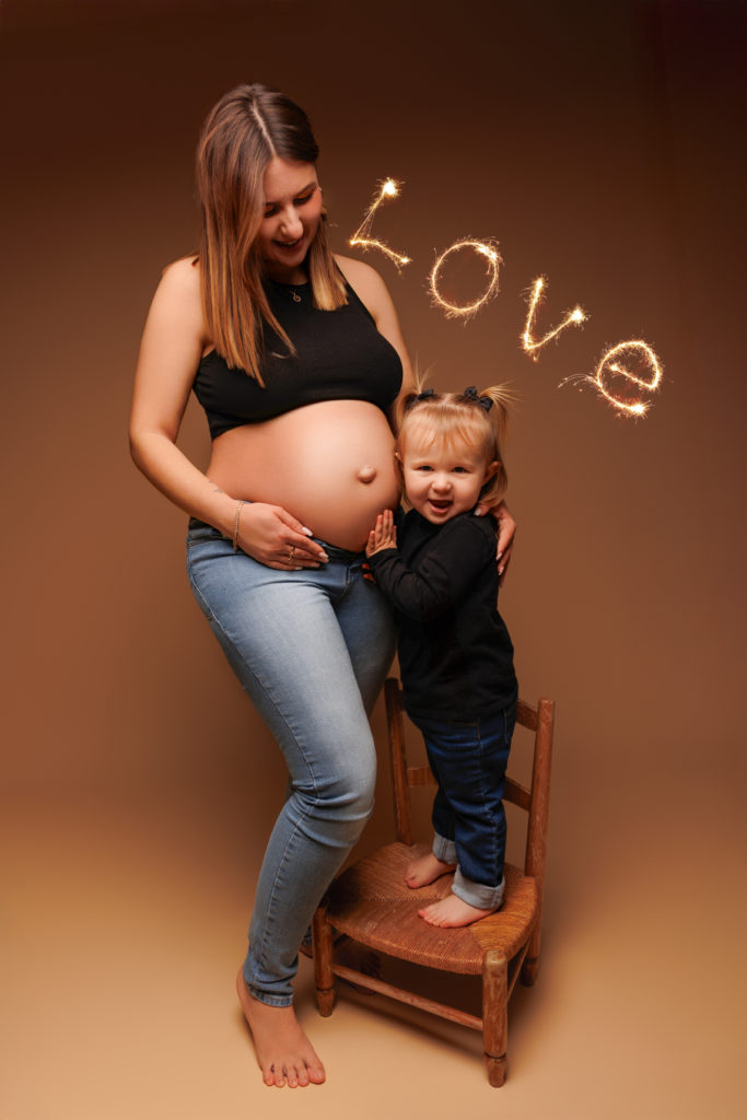 Femme enceinte souriante avec sa petite fille debout sur une chaise, posant main sur le ventre de sa maman, avec le mot "Love" écrit en lumière dans le fond