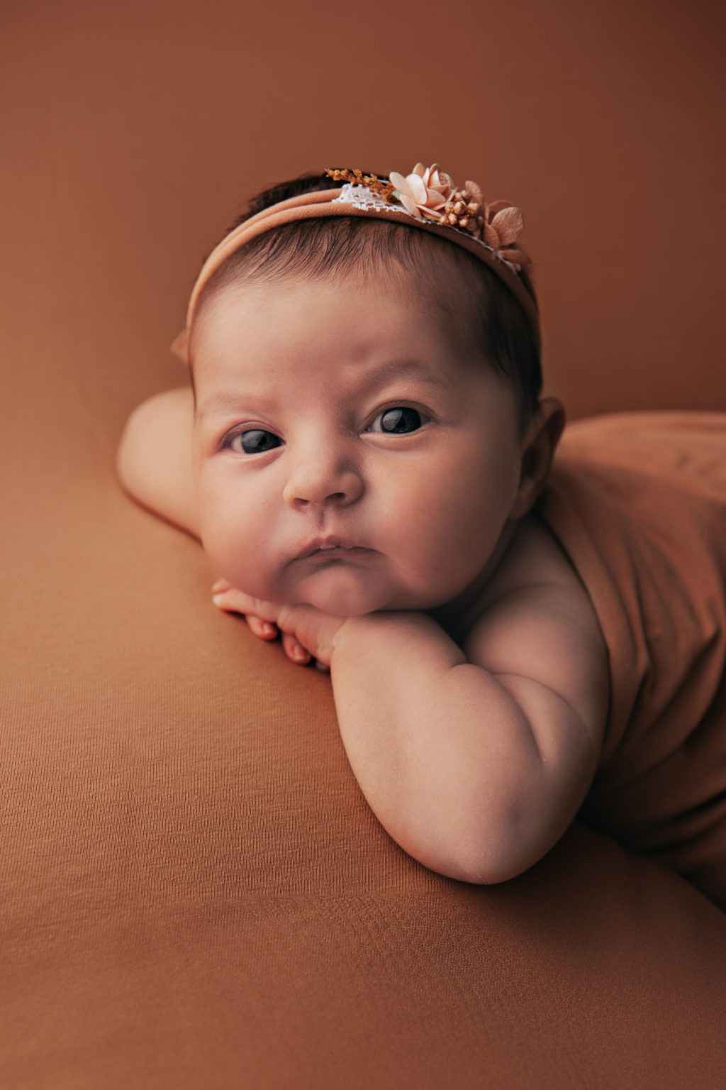 Portrait d’un bébé éveillé, allongé sur le ventre, regardant l’objectif avec intensité, bandeau fleuri sur la tête.