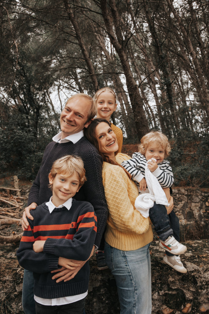 Portrait d’une famille de cinq personnes en extérieur : parents et trois enfants souriants, habillés en tenues automnales décontractées, dans un décor boisé naturel