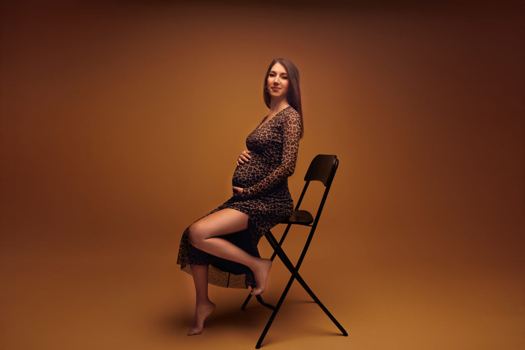Femme enceinte assise sur une chaise, en robe moulante léopard, dans une ambiance chaleureuse et élégante