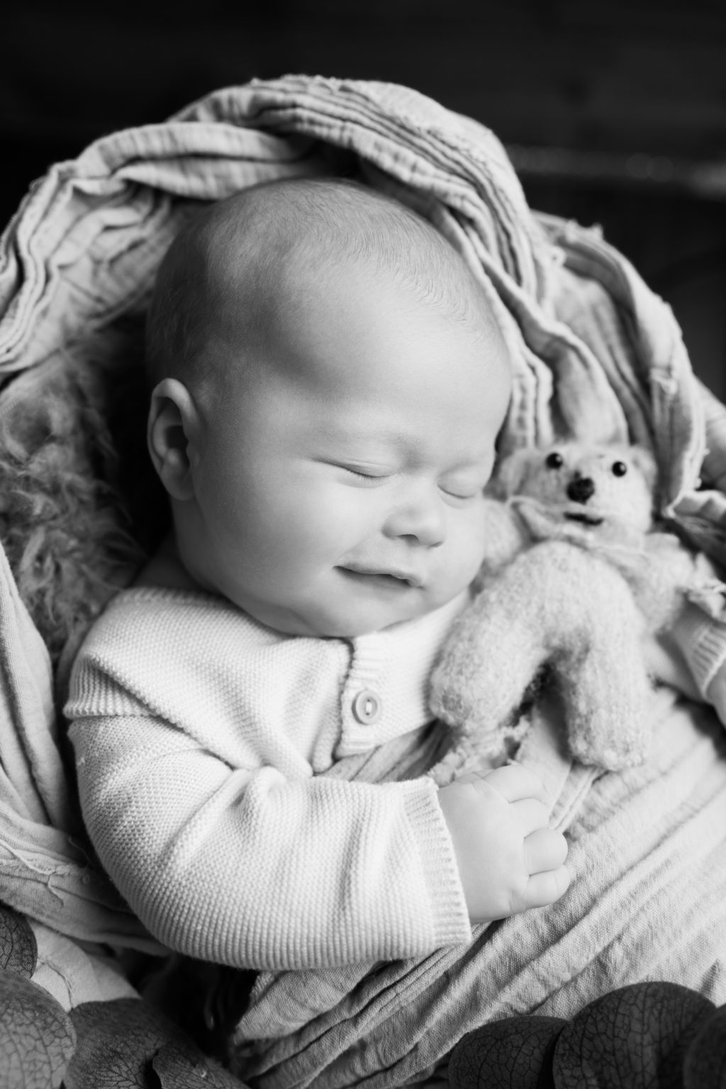 Bébé endormi souriant, en noir et blanc, avec un doudou dans les bras.