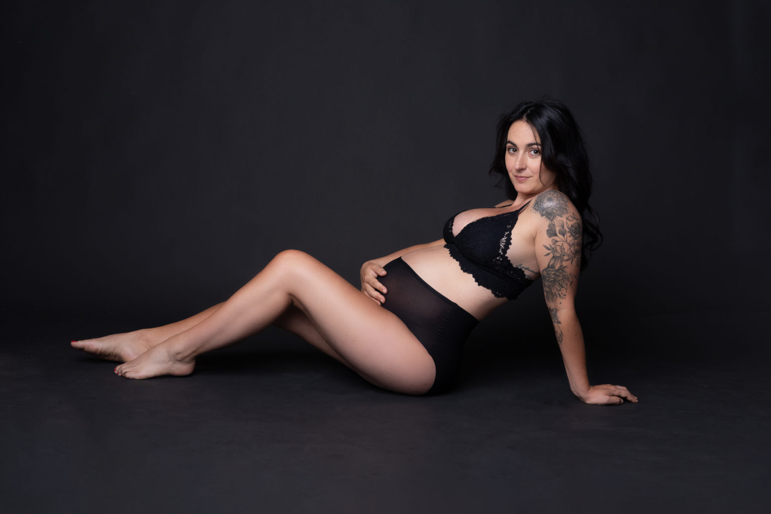 Femme enceinte assise au sol en lingerie noire, posant avec grâce et assurance devant un fond noir.