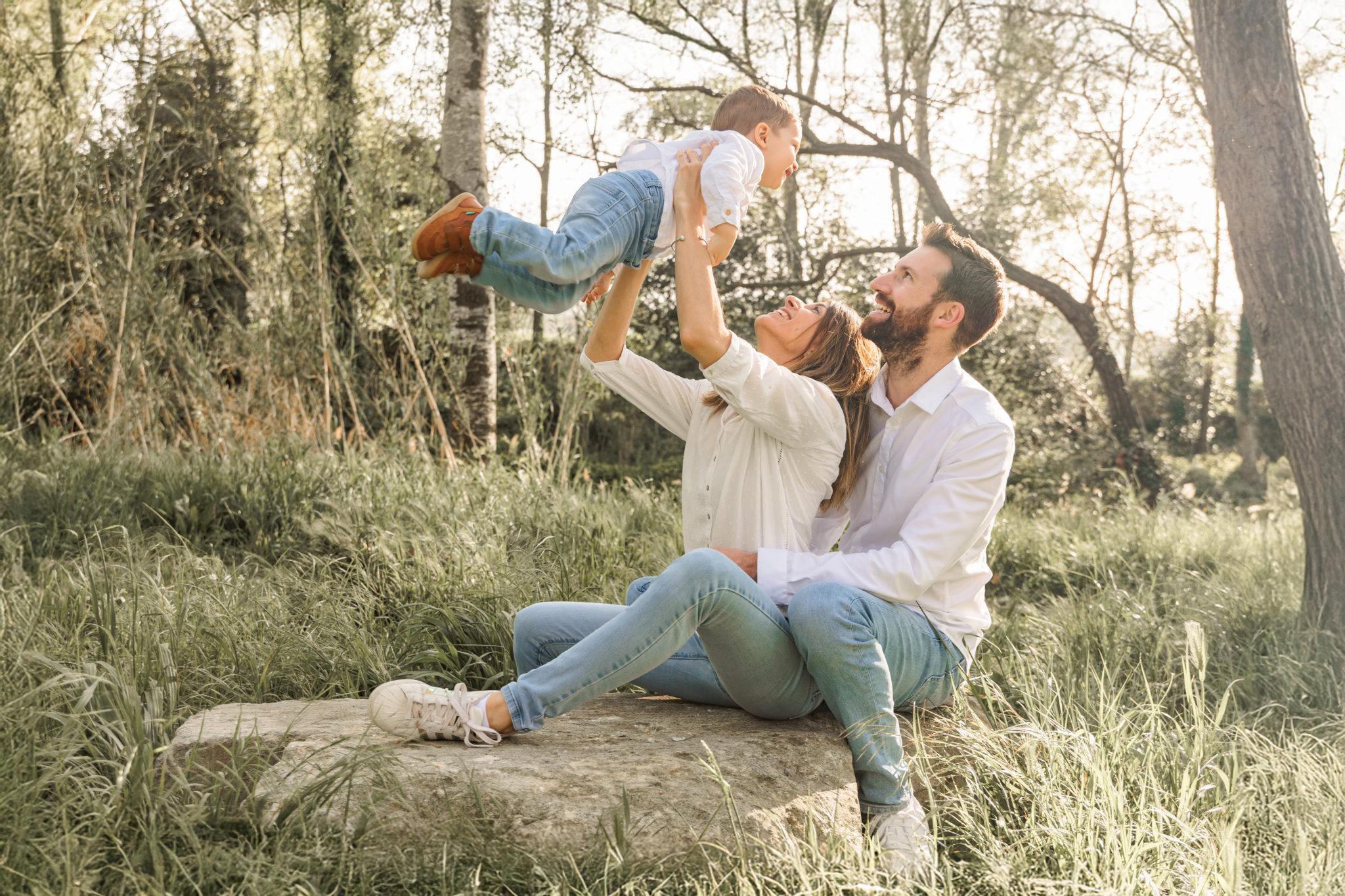 Famille heureuse en pleine nature : les parents assis sur une pierre regardent leur jeune enfant être soulevé en riant par sa maman