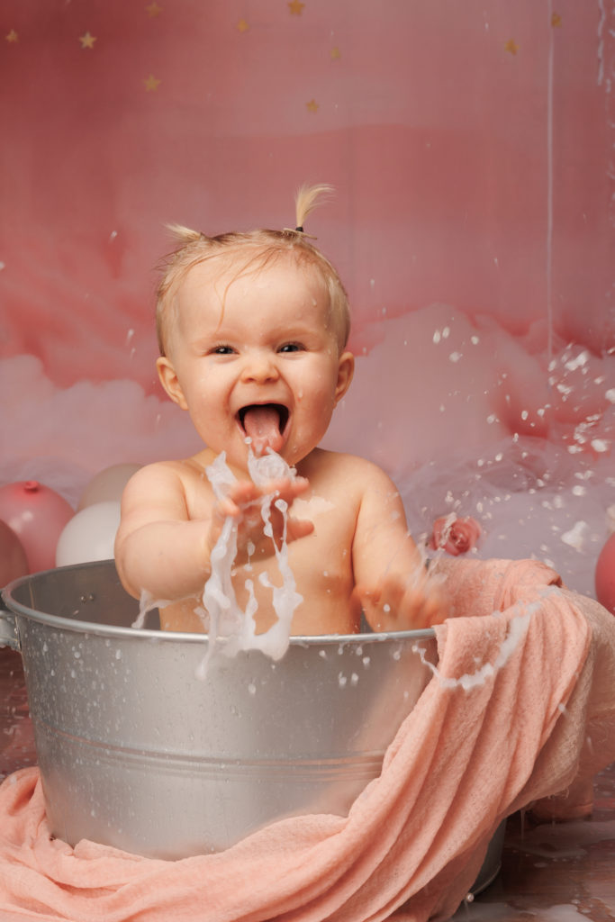 Bébé joyeux dans une bassine de bain en métal, éclaboussant de l’eau et riant à pleine bouche, sur un décor rose avec ballons et tissu doux
