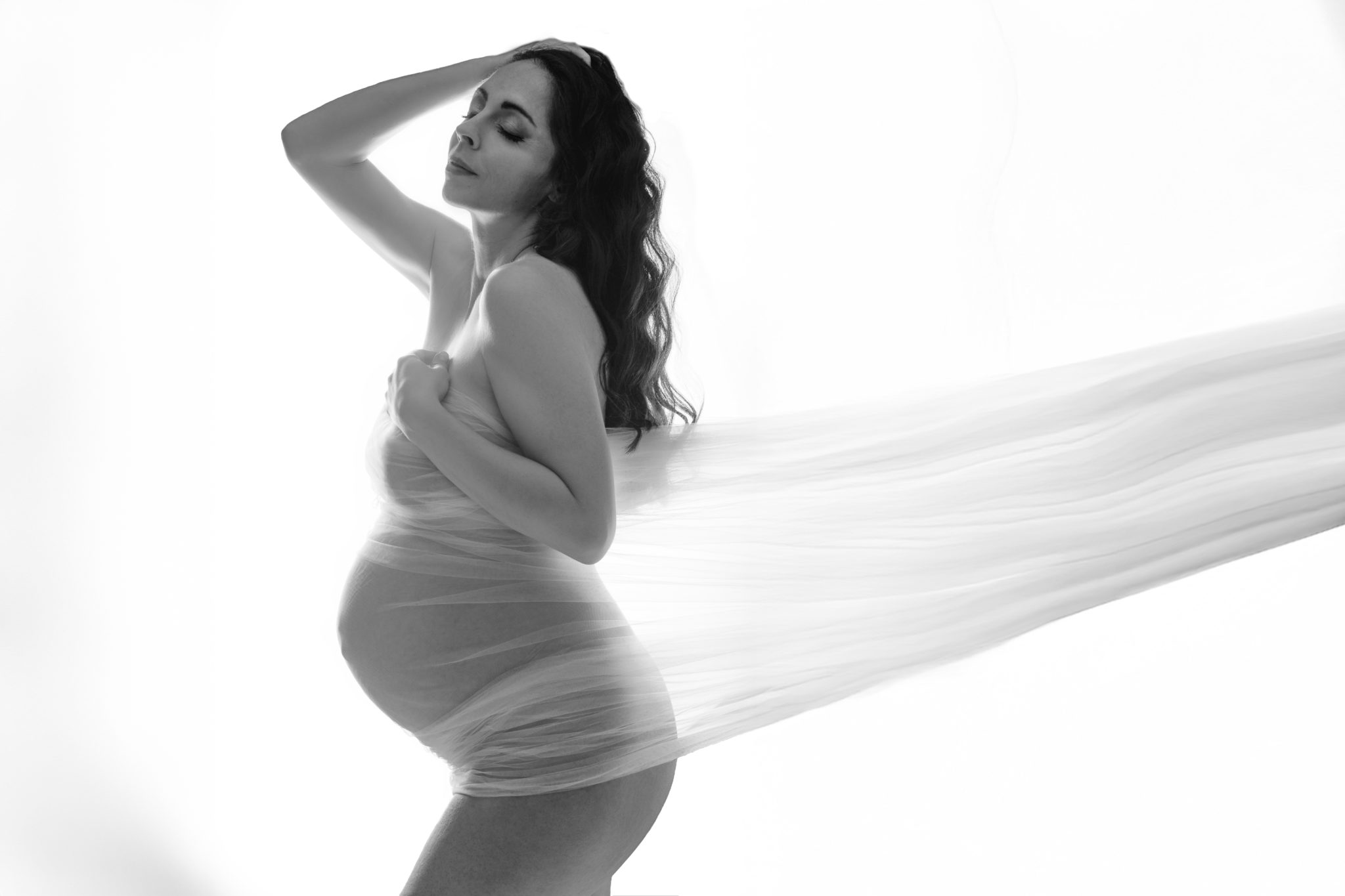 Femme enceinte en studio, enveloppée dans un voile fluide et lumineux, posant avec grâce sur fond blanc.