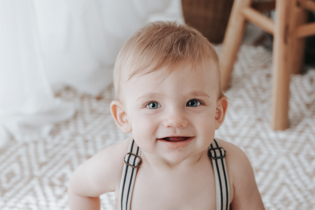 Bébé blond souriant portant des bretelles rayées, assis sur un tapis aux motifs géométriques dans une ambiance naturelle et lumineuse.