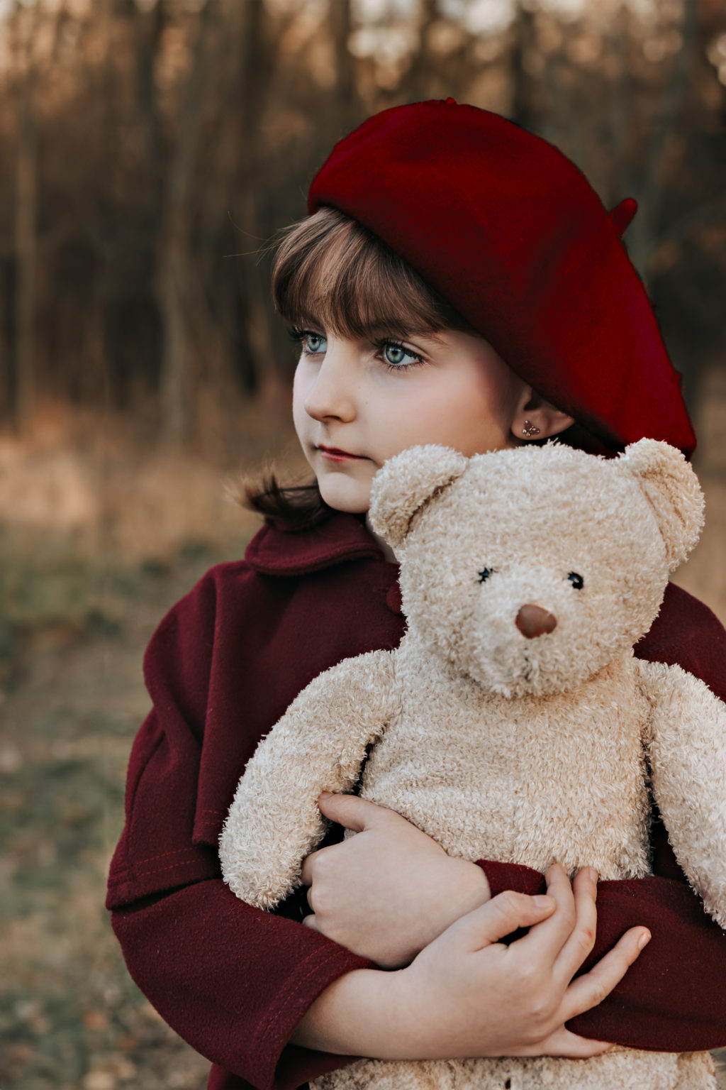 Petite fille en manteau bordeaux serrant son ours en peluche contre elle, regard rêveur vers l’horizon