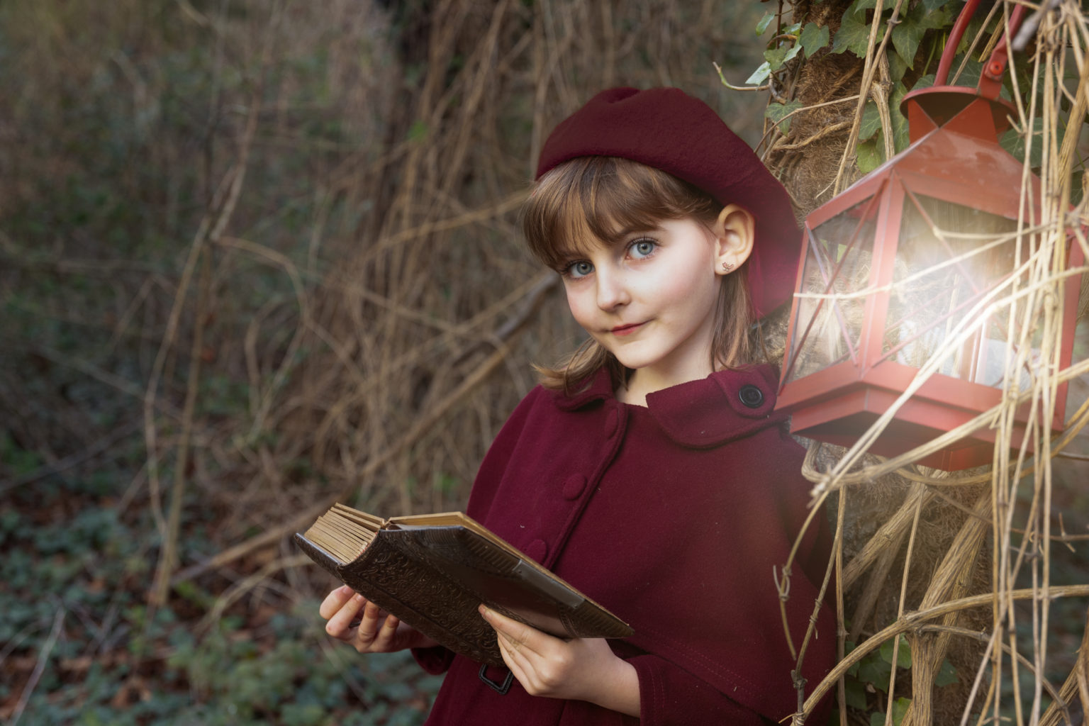 Petite fille en manteau rouge tenant un vieux livre à la lumière d’une lanterne, dans un décor naturel féérique