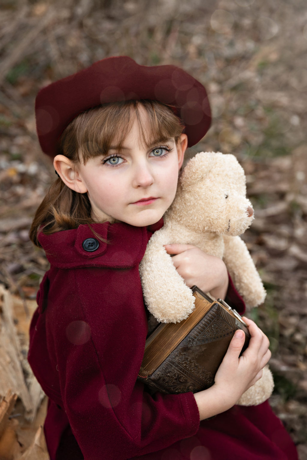 Petite fille en manteau bordeaux tenant un ours en peluche et un livre ancien, dans un décor naturel d’automne