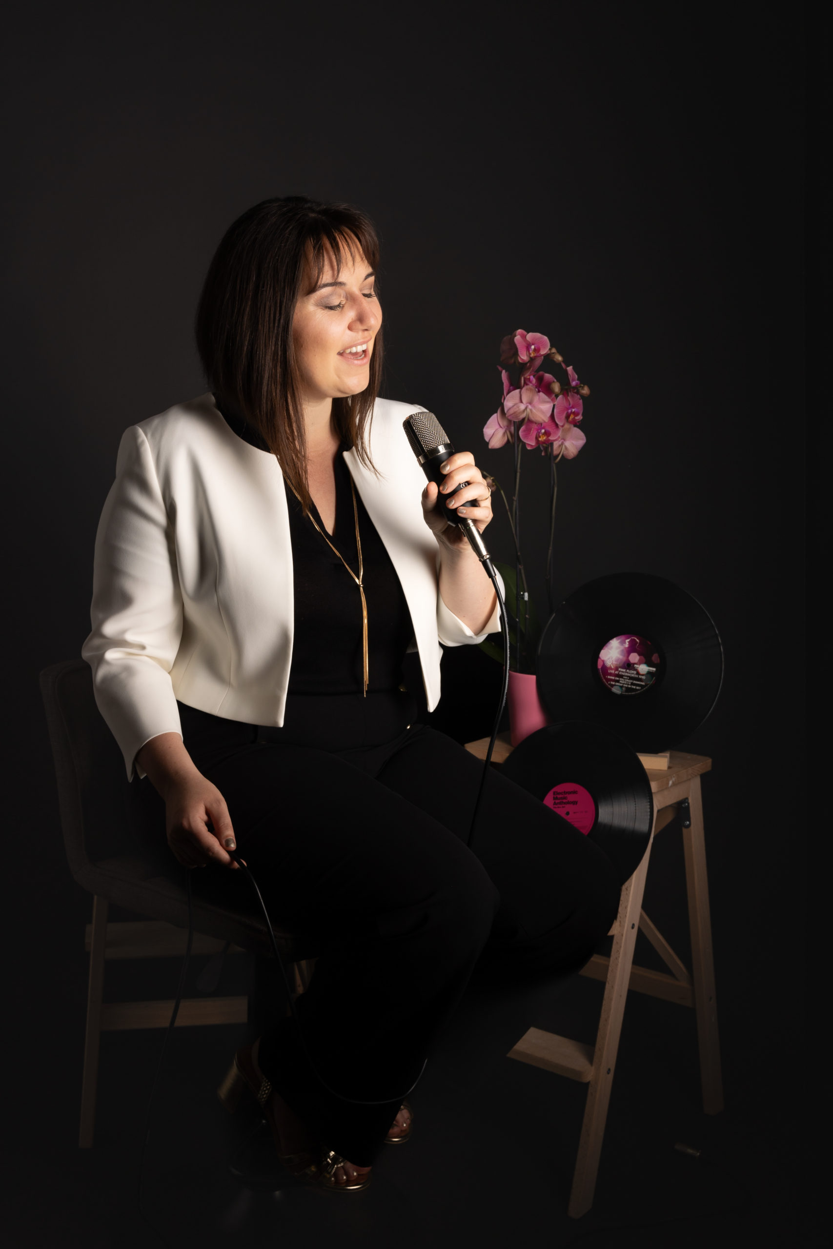Portrait en studio d’une femme élégante tenant un micro, assise près d’une orchidée et de disques vinyles, sur fond noir