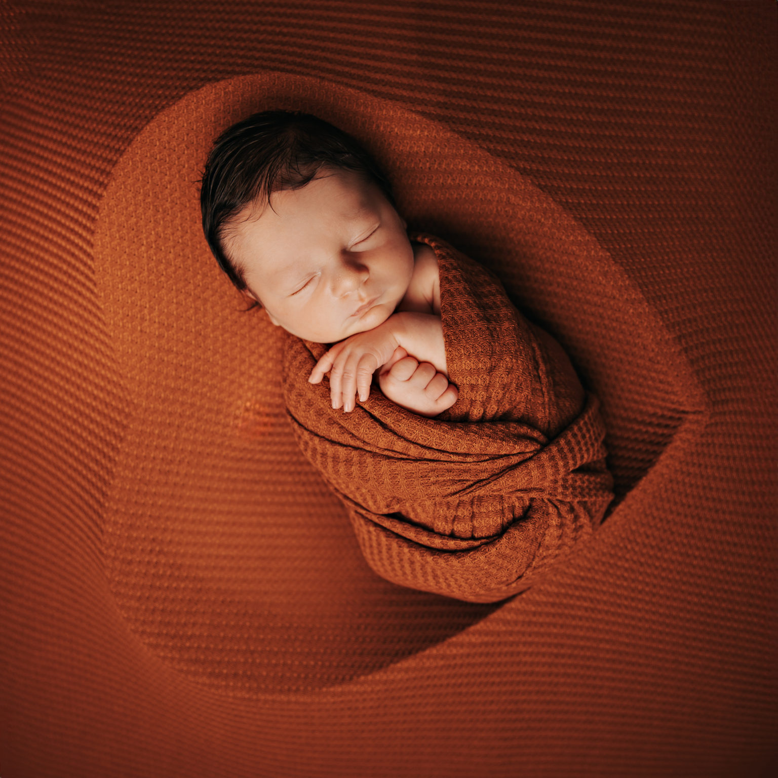 Nouveau-né emmailloté dans des tissus terracotta, allongé sur une surface assortie en forme de cœur