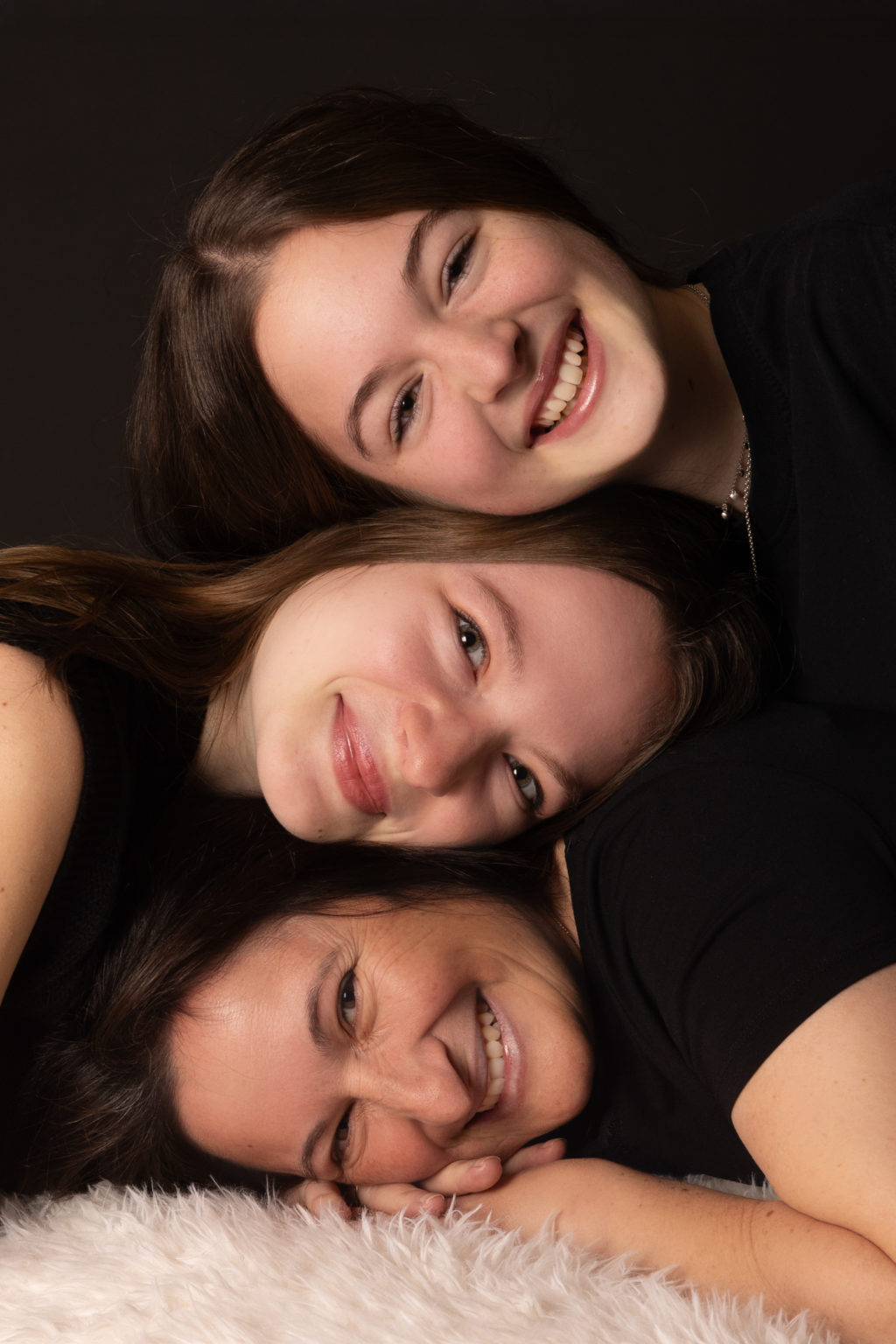 Portrait studio d’une maman et de ses deux grandes filles allongées les unes sur les autres, souriantes et complices, sur fond noir avec tapis blanc
