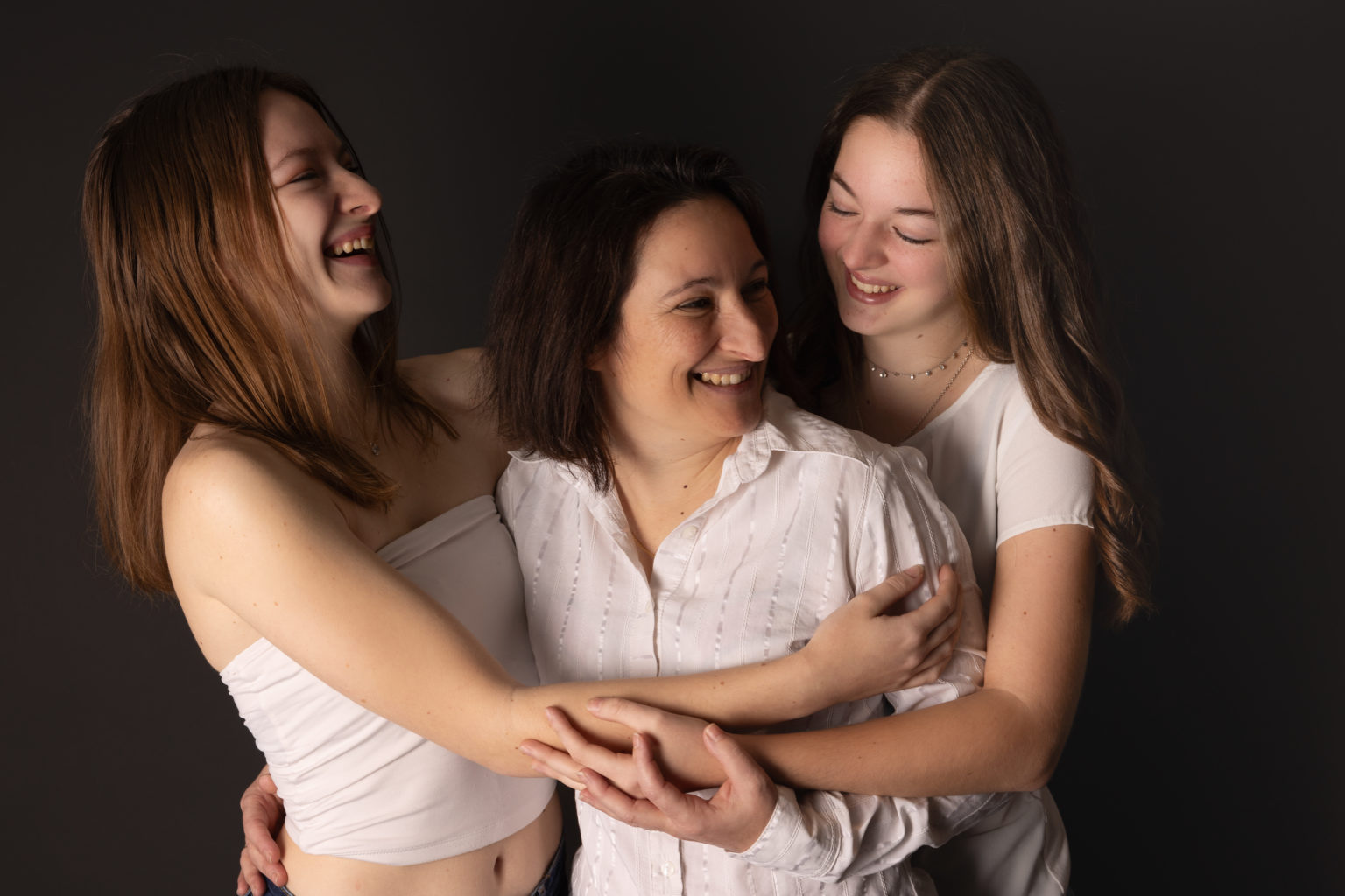 Maman entourée de ses deux filles, toutes les trois souriantes, dans un moment de tendresse et de complicité sur fond studio noir.