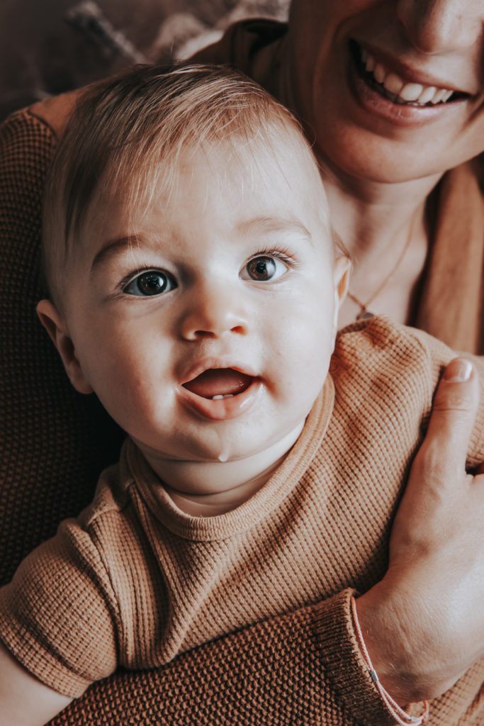 Portrait d’un bébé aux yeux clairs, vêtu d’un haut en maille beige, dans les bras souriants de sa maman
