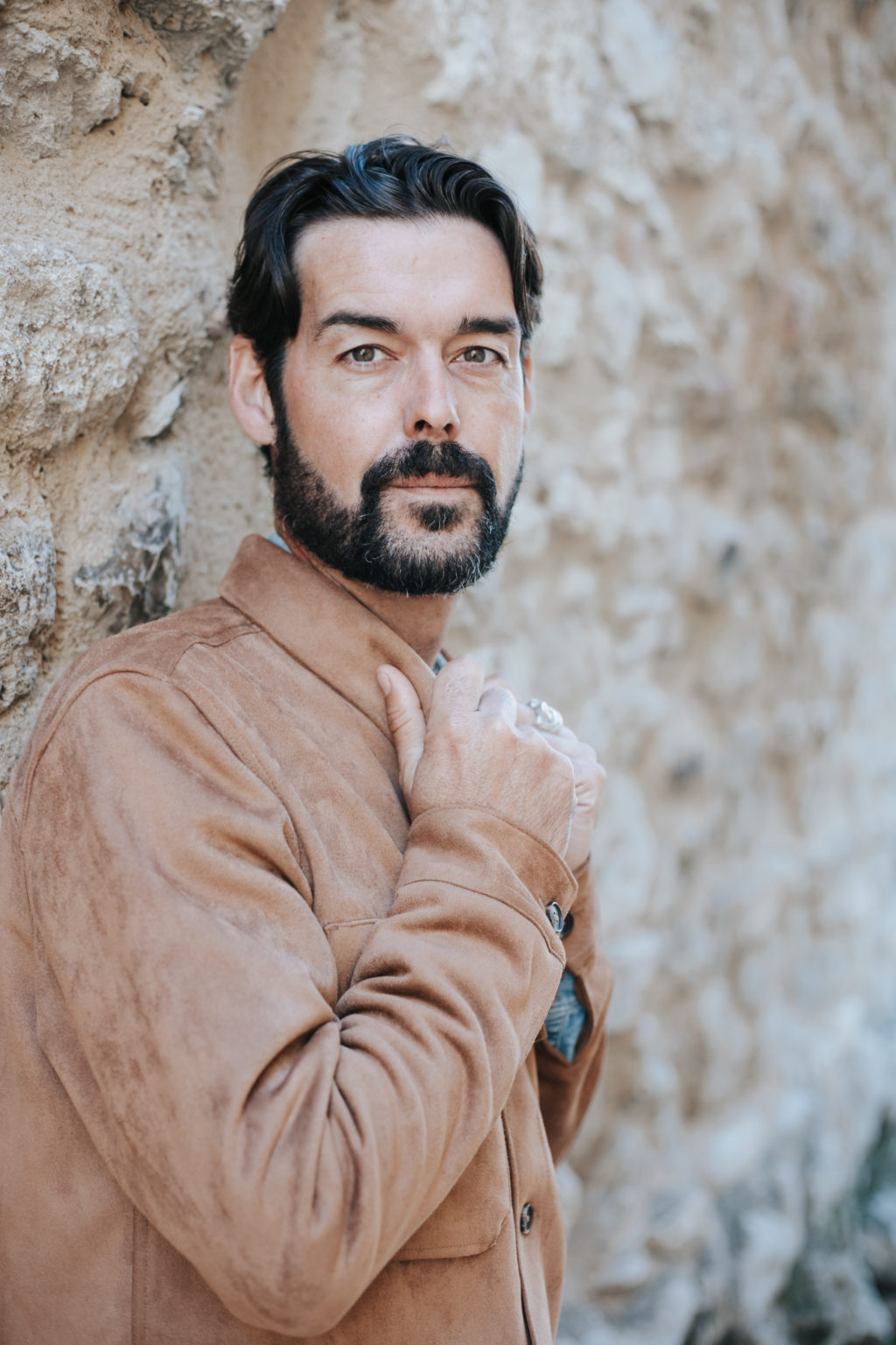 Portrait d’un homme barbu adossé à un mur en pierre, regard direct vers l’objectif