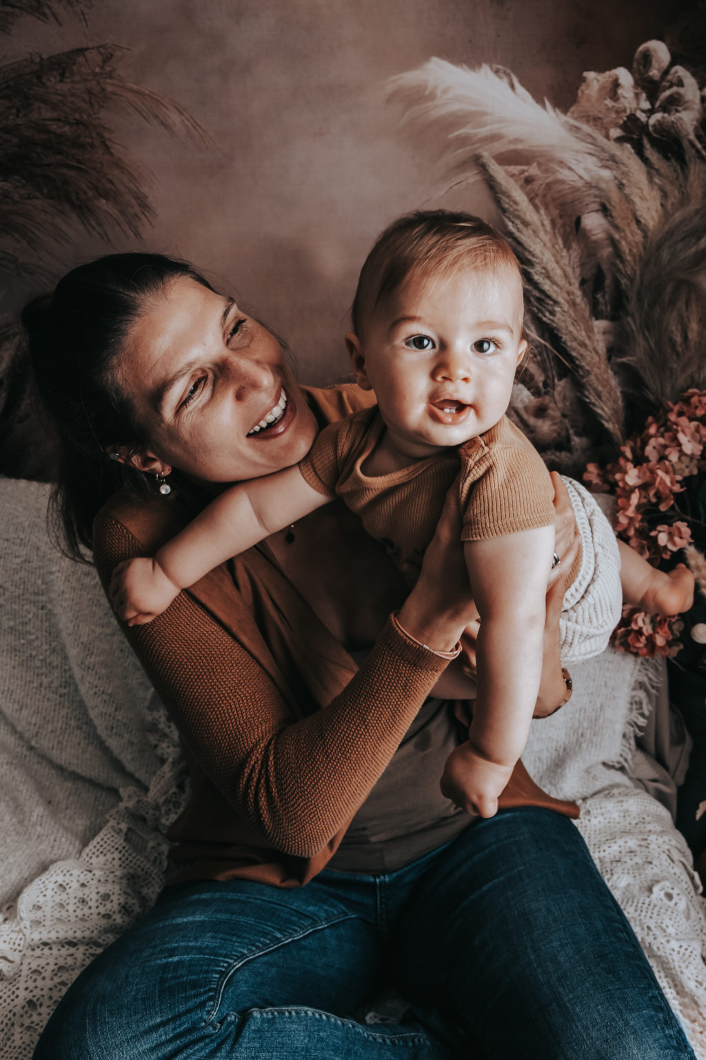 Maman souriante tenant son bébé dans les bras, moment complice sur fond naturel